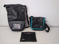 Photo showing the front view of all three items: large black waterproof Faraday tote with clear window, small black Faraday bag with blue piping, and black messenger bag with turquoise trim.
