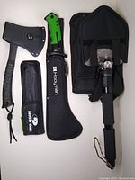 Front view of Schrade hatchet with sheath and Mossy Oak saw sheath, Hooyman tool sheath, and FiveJoy folding shovel in nylon case, showing handles and protective covers.