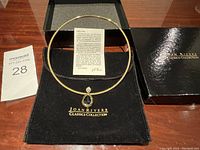 Gold-tone Joan Rivers Classics Collection necklace with teardrop blue stone pendant displayed on black pouch with certificate card and original box.