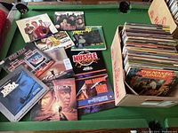 Photo showing a cardboard box filled with numerous Laser Discs, and several Laser Disc covers laid out on a pool table including titles such as Star Trek, Muscle Shoals, Van Damme Hard Target, and The Naked Jungle.