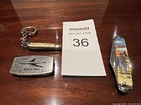 Photo of three items from lot: metal money clip with airplane engraving, small keychain folding knife and medium folding knife with horse rider illustration on handle.