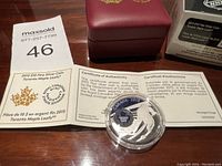 2015 Silver Coin in capsule, Certificate of Authenticity, maroon presentation box, and numbered MaxSold tag on wood table