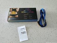 EMS muscle stimulator box with image of woman’s abdomen and yellow electrode pads shown on front, along with device features and blue/black wires attached.