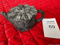 Black clay teapot shown from top and side angles on red quilted fabric background, highlighting leaf details and flower bud lid handle.