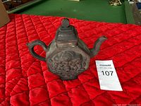 Front and side view of vintage hexagonal purple clay teapot with floral embossed design, handle, spout, and lid on a red quilted surface with MaxSold tag number 107.