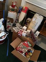 Photo of assorted Christmas decorations including boxed ornaments, potted trees, and lawn stakes arranged near a fireplace.