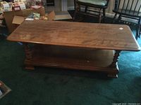 Front and side view of rectangular wooden coffee table on green carpet, showing top surface and turned legs with lower shelf.