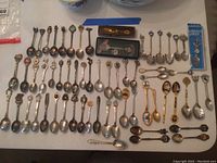 Full display of 47 collectible spoons arranged on a table, including pewter, gold plated, silver plated spoons in various designs and sizes. Three spoons appear in boxes with one blue boxed pewter spoon clearly visible.