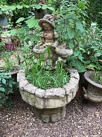 Full view of weathered concrete fountain in a garden setting surrounded by plants and dirt in basin.