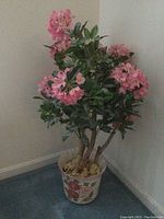 Full view of faux plant with pink flowers and green leaves in floral ceramic planter on carpeted floor near corner wall.