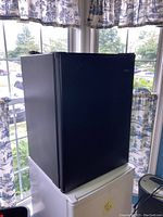 Front view of black Haier mini fridge with door closed, showing brand logo, situated indoors near window with patterned curtains.