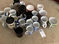 Overall view of 23 coffee mugs arranged on carpet, including 3 travel mugs and various ceramic mugs