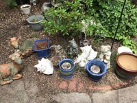 Photo showing outdoor garden setting with two concrete deer figurines, ceramic planters, several animal figurines and large white conch shells arranged on soil and mulch near some bushes