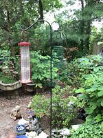 Black metal shepherds hook with a red and white cylindrical mesh bird feeder hanging, surrounded by garden plants and statues.