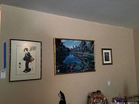 A wall section displaying three framed wall art pieces. From left to right: a framed geisha artwork, a central blue and green Hawaiian river scene with a boat, and a smaller framed black and white print.