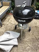 Black Weber charcoal kettle grill on tripod stand with two wheels and handle. Grill lid has vent and metallic handle. The grill is placed on a stone patio with some garden furniture visible.