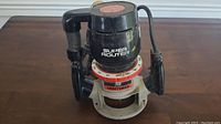 Photo showing Craftsman Super Router power tool standing on a wooden surface, black body with red adjustable depth ring and handles.