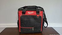 Front view of Milwaukee jobsite tech bag showing red and black colors, multiple compartments, carry handles, and shoulder strap