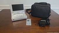 Photo of silver Polaroid portable DVD player with screen open, remote control, power adapter, and black carrying case on wooden surface.
