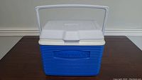 Front view of small Rubbermaid cooler showing white lid and handle, ribbed blue body with embossed brand name.