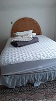 Full bed setup showing queen size mattress with quilted white cover, light blue skirt on box spring, rattan curved headboard, pillows, and folded comforter on bed.