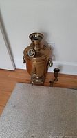 Full view of vintage brass samovar on floor with brass medallion hanging from chimney, small shot cup and vase nearby