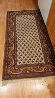 Full view of one carpet showing central cream field with repeating dark motifs and multi-colored border in brown and reddish tones, placed on wood floor.