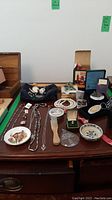 Overview of table top showing wooden box, black pouch with silver cases, jewellery, decorative plate, lighter, clock box and wooden roller tool