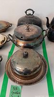 Three tarnished silverplate covered serving bowls, two with round knob handles and one cloche with beaded loop handle, arranged on green tape for size reference