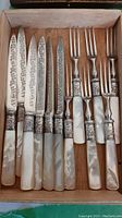Full view of six knives with mother of pearl handles and etched blades alongside six matching forks in a wooden box.