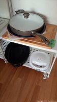 Shelf arrangement showing frying pan with lid on wooden cutting board above black roasting pan and white waffle pan. Also shows plastic cutting board beside frying pan.