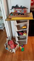 Photo showing six stackable plastic box shelves holding various toys including dinosaur models, metal tin box, toy cars and train engine. Also includes toys and boxes on the floor nearby.