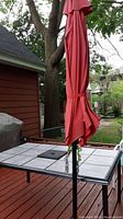 Full view of patio table with fifteen white/light gray tiles in black metal frame and closed red umbrella inserted in center.