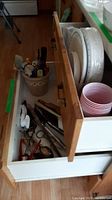 Two open kitchen drawers showing contents of utensils, plastic storage savers, paper plates, and bowls.