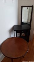 Black side table with drawer and lower shelf beside the round wooden side table, both showing scratches and wear.