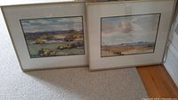Both framed prints leaning against wall on carpeted floor