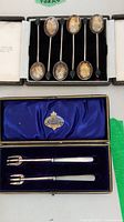 Top view of six tarnished silver tea spoons with black decorative ends in black velvet case; two pickle forks with white handles in blue satin lined case.