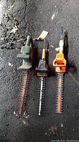 Three electric hedge trimmers laid on floor, showing worn and dirty condition of each unit.