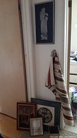 Full view of material wall hanging draped, framed wall pictures leaning on wall including medieval knight figure, and small waste basket partly visible.