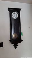 Full front view of wooden wall clock with pendulum and Roman numeral dial.