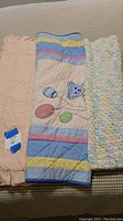 Photo showing all three blankets: peach eyelet quilt with ruffle, homemade quilt with teddy bear motif and red gingham reverse, and crocheted blanket with blue, pink, and yellow yarns.