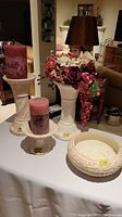 Four antiqued ceramic items displayed on a table: two candle holders each with a pink candle, an antiqued ceramic stand with a flower and fruit arrangement, and a decorative carved bowl.