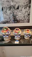 Front view of three Fitz and Floyd canisters shaped like fruit baskets of varying sizes on a black surface under artwork.