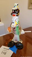 Front view of a colorful blown glass clown figurine with orange ball in hand and cigar in other hand, seated on blue tape with auction number 116.