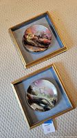 Two custom framed decorative plates painted by Thomas Kinkade placed on floor carpet, showing gold-tone frames and light blue backing.