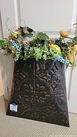 Full view of embossed black metal container holding artificial flowers including yellow and cream roses, green ivy leaves, and small blue berry clusters.
