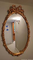 Full view of oval mirror hung on wall showing gold floral and bow decorative frame.
