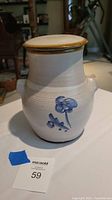 Front and side view of a stoneware pottery jar with blue hand-painted floral decoration and small side handles on an off-white surface.