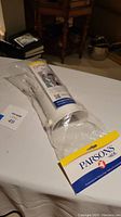Photo of the Parsons sock aid in original packaging showing front side with brand and product name, clear plastic sleeve around white rigid plastic device with yellow strip handle.