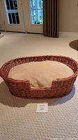 Wicker pet bed with tan cushion in carpeted room, showing side angle with basket weave details and shape.